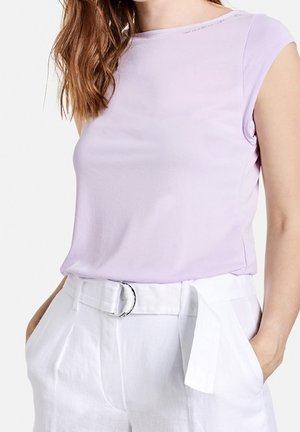 Frau trägt ein hellviolettes ärmelloses Top, das in hoch taillierte weiße Hose mit passendem Stoffgürtel gesteckt ist, und hat die Hände in den Taschen.