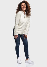 Veste en polaire blanche avec un col haut, surface texturée ; associée à un jean skinny noir et des baskets blanches. Le modèle se tient dans une pose ludique.