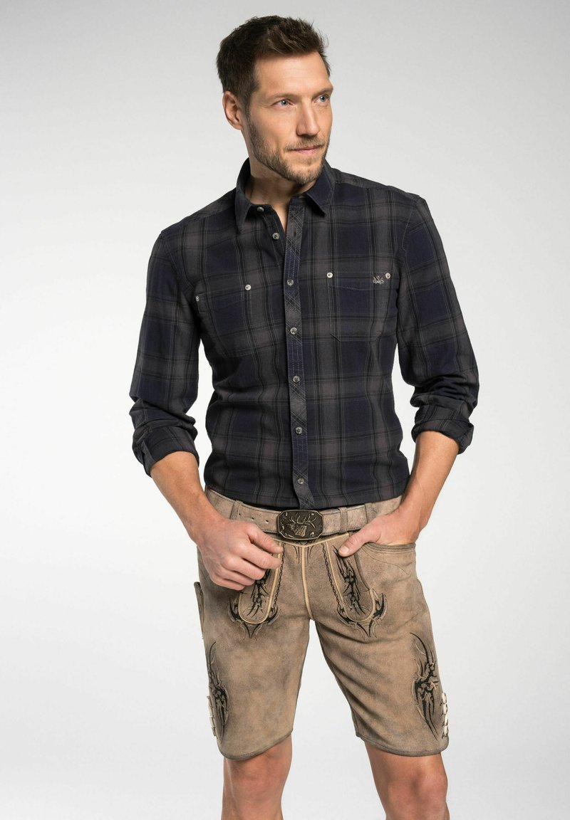 Marineblaues kariertes Langarmhemd mit Knopfdetails, kombiniert mit beigen Ledershorts, die aufwendige Stickereien und eine dekorative Schnalle aufweisen.