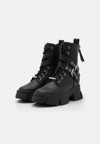 Botas de tobillo negras gruesas con cordones, suelas gruesas, doble correa y detalles de hebilla plateada, y un tirador de cremallera en la parte posterior.