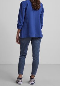 Blazer bleu à coupe ajustée, manches retroussées et tissu texturé, associé à un jean bleu foncé et des baskets multicolores.