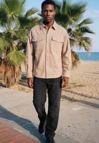 Homme marchant sur le trottoir au bord de la plage, portant une chemise beige à boutons et un jean noir, avec des palmiers et l’océan en arrière-plan.