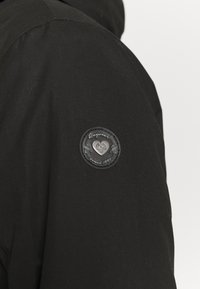 Veste noire avec une épaule présentant un patch circulaire noir avec un logo embossé et du texte "Raquwear depuis 1997." Texture lisse et mate.