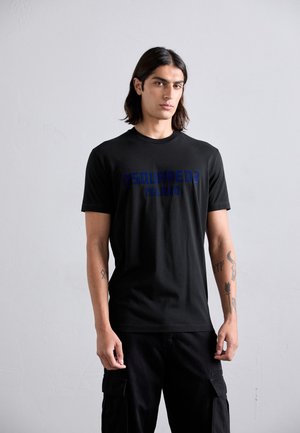 Young man with long dark hair wearing a black DSQUARED2 Milano t-shirt and black cargo pants, standing against a plain gray wall.