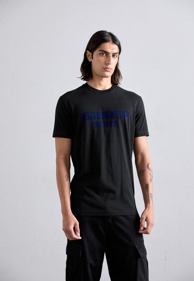 Jeune homme avec de longs cheveux bruns, portant un t-shirt noir DSQUARED2 Milano et un pantalon cargo noir, appuyé contre un mur gris uni.