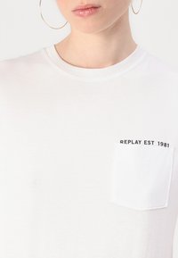 White cotton T-shirt featuring a round neck, short sleeves, and a left chest pocket with "REPLAY EST 1981" printed in black.