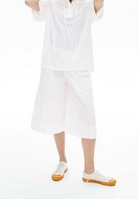 Vit kortärmad topp med dragsko i kragen, kombinerad med vita knälånga shorts och vita sneakers med orange detaljer.