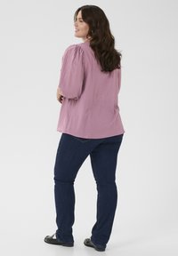 Kvinde med langt brunt hår iført en løs lyserød bluse, mørkeblå jeans og sorte sko, stående og kiggende til siden.