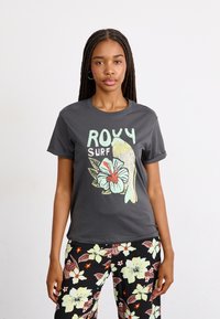 T-shirt en coton gris foncé avec des manches retroussées, arborant un graphisme sur le thème du surf dans des couleurs pastel, associé à un pantalon noir à motifs floraux.