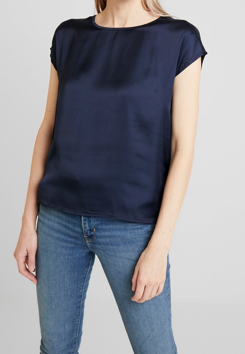 Kvinde iført en kortærmet marineblå satinbluse og blå denimjeans foran en ensfarvet lys grå baggrund.