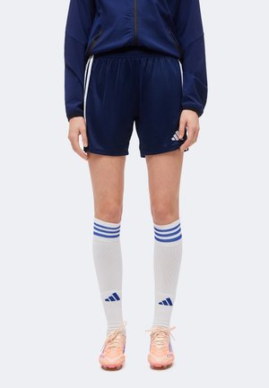 Person iført marineblå atletikshorts, hvide knæstrømper med blå striber, ferskenfarvede fodbolddsko og en marineblå jakke.