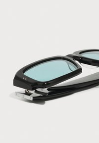 Gafas de sol negras con montura rectangular, lentes tintados de azul y un detalle cromado en la patilla con iniciales grabadas.