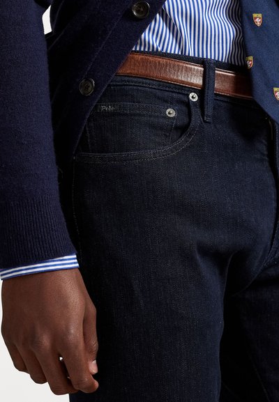 Hombre vistiendo jeans de mezclilla azul oscuro, cinturón de cuero marrón, camisa a rayas azul y blazer azul marino con parches de escudo bordados.