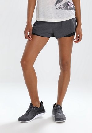 Ben af en person iført grå sportsshorts med sort kant, en hvid mønstret top og grå sneakers med hvide såler.