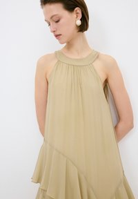 Helles beige ärmelloses Kleid mit einem plissierten Ausschnitt und ausgestellt geschnittenem Saum. Weicher, fließender Stoff mit drapiertem Design. Goldene Ohrringe sichtbar.