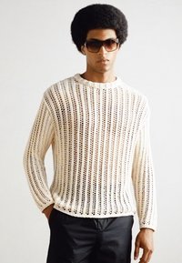 Suéter de punto color crema con un patrón texturado abierto. Mangas largas y cuello redondo. Llevado con pantalones cortos oscuros. El modelo lleva gafas de sol.