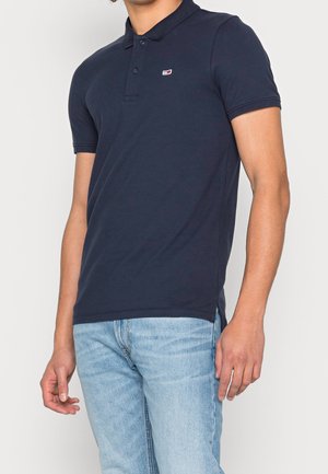 Man in een marineblauwe polo met korte mouwen en lichtblauwe jeans, staande tegen een effen witte achtergrond.