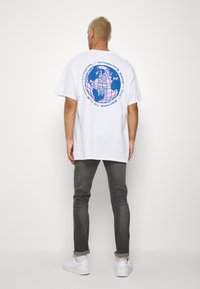 Hombre con el pelo rubio corto, vistiendo una camiseta gráfica blanca, jeans oscuros arremangados y zapatillas blancas, de pie frente a un fondo sencillo.
