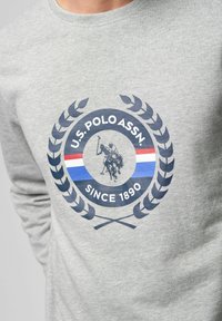 Grijze langslobige shirt met een cirkelvormig logo van een polo-speler, laurierbladeren en de tekst "U.S. POLO ASSN. SINCE 1890" in donkerblauwe en rode strepen.