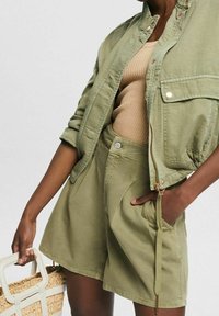 Vihreä khaki-takkia, jossa on kaulus ja taskut, yhdistettynä samanvärisiin shortseihin. Asusteena on teksturoitu beige paita ja kudottu korikassi.