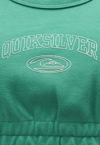 Tissu vert côtelé avec le texte "QUIKSILVER" en blanc souligné, formant un arc au-dessus du logo de la marque représentant une vague et une montagne en contour blanc.