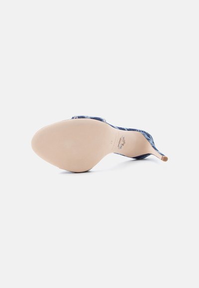 Γυναικείο μπλε παπούτσι με ψηλό τακούνι ξαπλωμένο στο πλάι, που δείχνει την λεία μπεζ σόλα και το λεπτό τακούνι πάνω σε απλό λευκό φόντο.