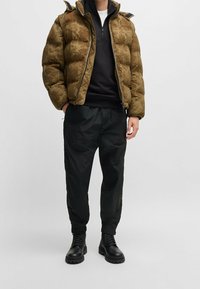 Puffer-takki teksturoidussa oliivinvihreässä, mustilla yksityiskohdilla, yhdistettynä mustiin kiristysnauhoilla varustettuihin housuihin ja tukeviin mustiin nauhakenkiin.