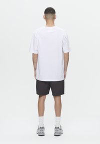 T-shirt en coton blanc, coupe oversize, assortie à un short noir. Modèle portant des baskets grises avec des chaussettes blanches, se tenant devant un fond uni.