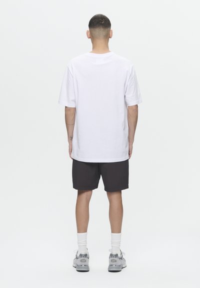 T-shirt en coton blanc, coupe oversize, assortie à un short noir. Modèle portant des baskets grises avec des chaussettes blanches, se tenant devant un fond uni.