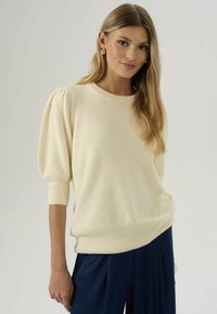 Jeune femme aux longs cheveux blonds portant un pull à manches bouffantes crème et un pantalon bleu marine, souriant sur un fond clair.