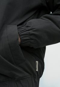 Melnā jaka ar elastīgajiem piedurknēm un sānu kabatu. Gluda, matēta auduma ar vieglu mirdzumu. Birka saka "Only & Sons."