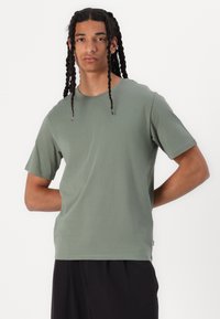 Tricou scurt cu mânecă scurtă de culoare verde măsliniu, dintr-un amestec de bumbac moale, având un decolteu elastic și o croială confortabilă, asortat cu pantaloni negri.