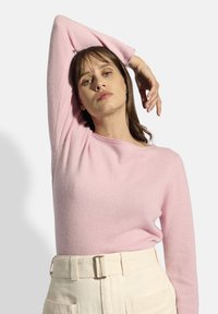 Helles rosa Strickpullover mit Rundhalsausschnitt und langen Ärmeln, kombiniert mit cremefarbenen, hoch taillierten Hosen, die einen Gürtel mit Schnalle und Taschen haben.