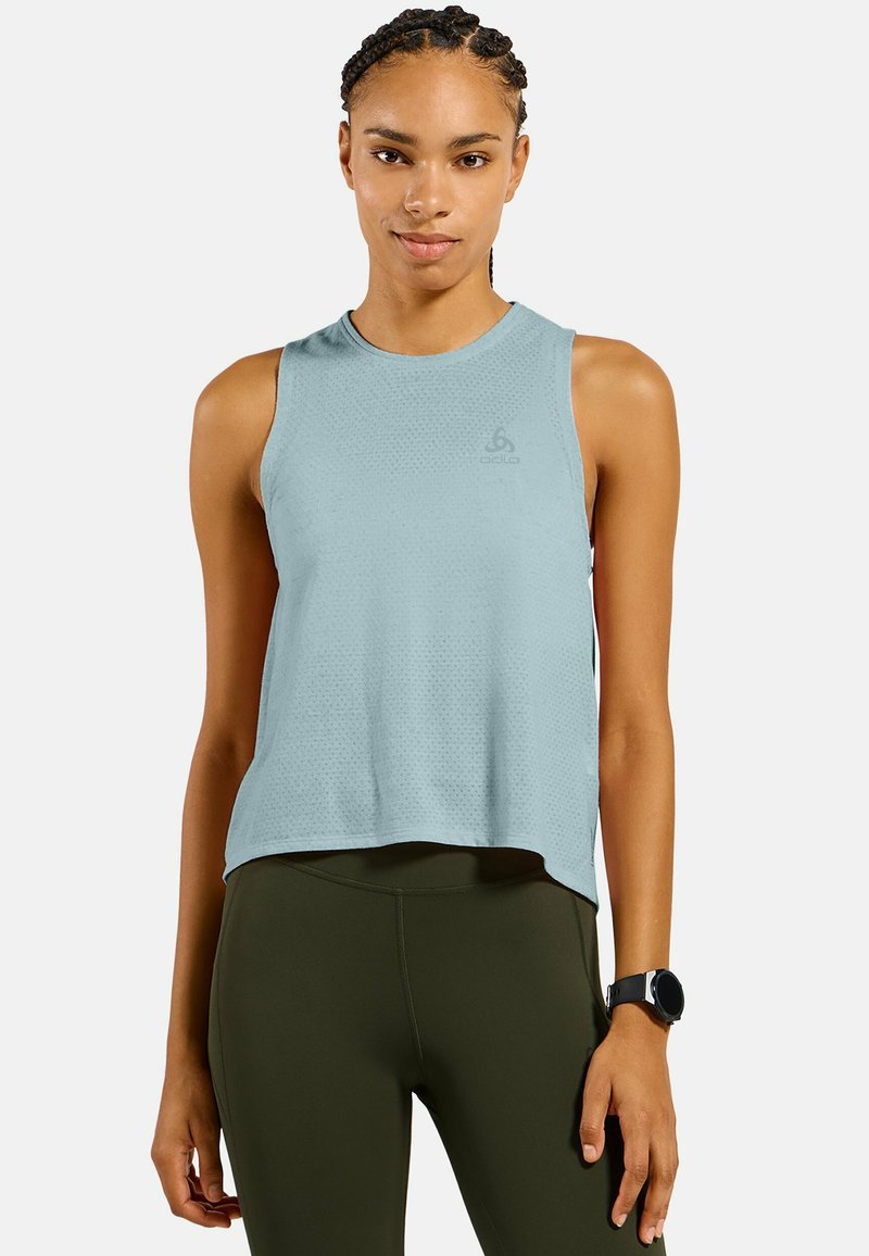 Junge Frau mit geflochtenem Haar, die ein hellblaues ärmelloses Sporttop und dunkelgrüne Leggings trägt, steht mit einer Smartwatch am Handgelenk.