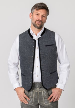Mann in traditioneller deutscher oder österreichischer Tracht mit weißem Hemd, dunkler strukturierter Weste und verzierten Lederhosen vor schlichtem Hintergrund.
