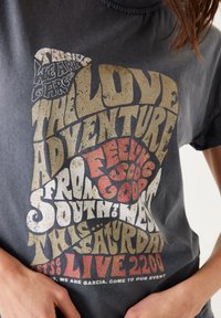 Mörkgrå bomullströja med djärv, flerfärgad retrotypografi som har text om ett evenemang. Sliten textur ger visuellt intresse.