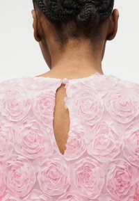 Οπίσθια όψη ατόμου με πλεξούδες που φορά ροζ φόρεμα με υφασμένα ροδοειδή σχέδια και μικρό κουμπί στο λαιμό.