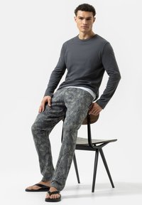 Grijze lange mouwen shirt, gebloemde blauwe en crèmekleurige broek, zwarte slippers; zittend op een stoel met houten en metalen elementen.