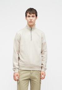 Junger Mann in einem beigen Quarter-Zip-Sweatshirt und hellen Khaki-Cargo-Hosen, der vor einem schlichten weißen Hintergrund steht.