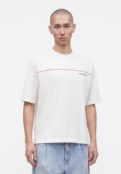 Homme à la tête rasée portant un t-shirt blanc à manches courtes avec une ligne rouge et un texte, associé à un jean ample bleu clair, sur un fond uni.