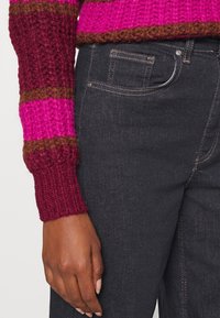 Stickad tröja med magenta, burgundy och bruna ränder; tillsammans med mörk denimjeans. Närbild av ärm och handdetalj.