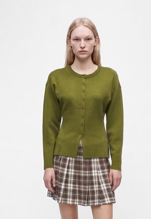 Jeune femme aux cheveux blonds et lisses portant un cardigan boutonné vert olive et une jupe plissée marron à carreaux, debout devant un fond uni.