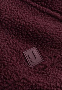 Burgundy fleece-kangas, jossa on teksturoitu pinta ja neliömäinen kumilogo, jossa on tyylitelty "J" alakulmassa.