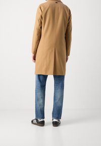 Cappotto beige in misto lana con taglio dritto, spacco alto sulla schiena e maniche lunghe, abbinato a jeans di denim blu e sneakers nere con strisce bianche.