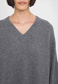 Personne portant un pull gris côtelé à col en V avec de courts cheveux foncés visibles du cou jusqu'au milieu de la poitrine sur un fond uni.