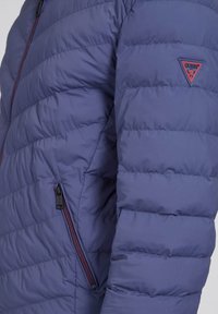 Veste matelassée bleu marine avec un motif de quilting horizontal, poche zippée et accents rouges, arborant un logo triangulaire sur la poitrine.