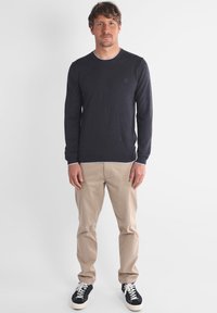 Homme se tenant contre un fond blanc, portant un pull à manches longues sombre, un pantalon beige et des baskets noires avec des lacets blancs.