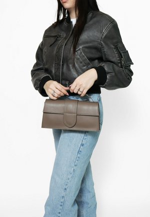 Grijze leren handtas met een klep-sluiting, rechthoekige vorm en gestikte accenten, vastgehouden door een persoon in een zwarte bomberjack en blauwe spijkerbroek.