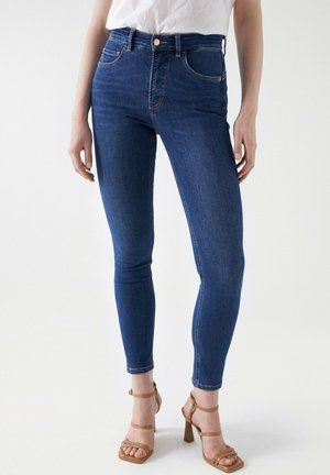 Jean skinny en denim bleu foncé avec taille haute, cinq poches et détails de couture orange, associés à des sandales à talons hauts à lanières beiges.