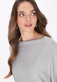 Lys grå, ribbet off-shoulder top med subtil glans. Lang brunt hår og perle-dråbe øreringe fuldender looket.